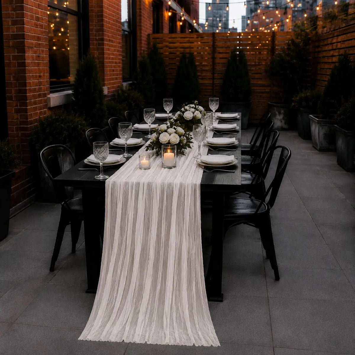 Chemin de table voilage plissé blanc sur table dressée en terrasse extérieure, avec guirlandes lumineuses et bouquet de roses