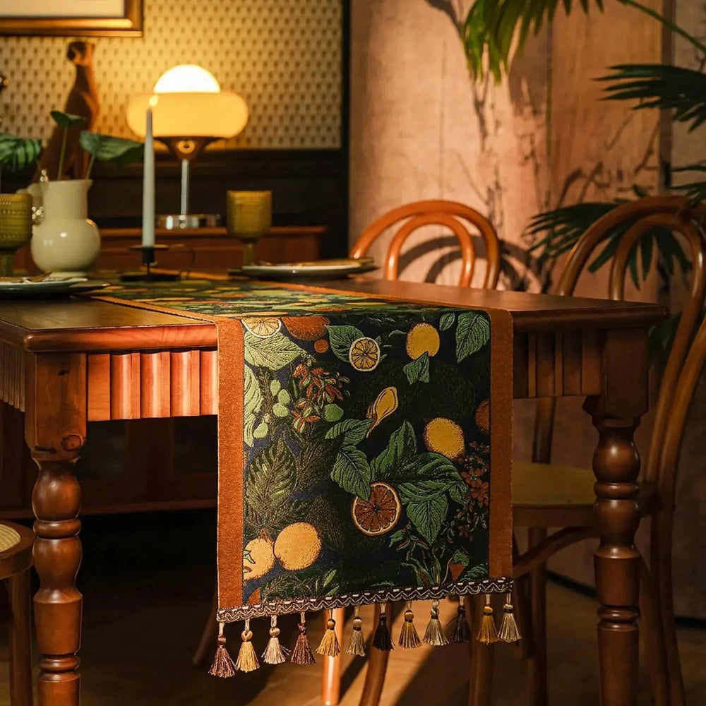Image d'un élégant chemin de table ayant des agrumes et des feuilles vertes comme motif, placé sur une table en bois brun avec lampe et décorations.