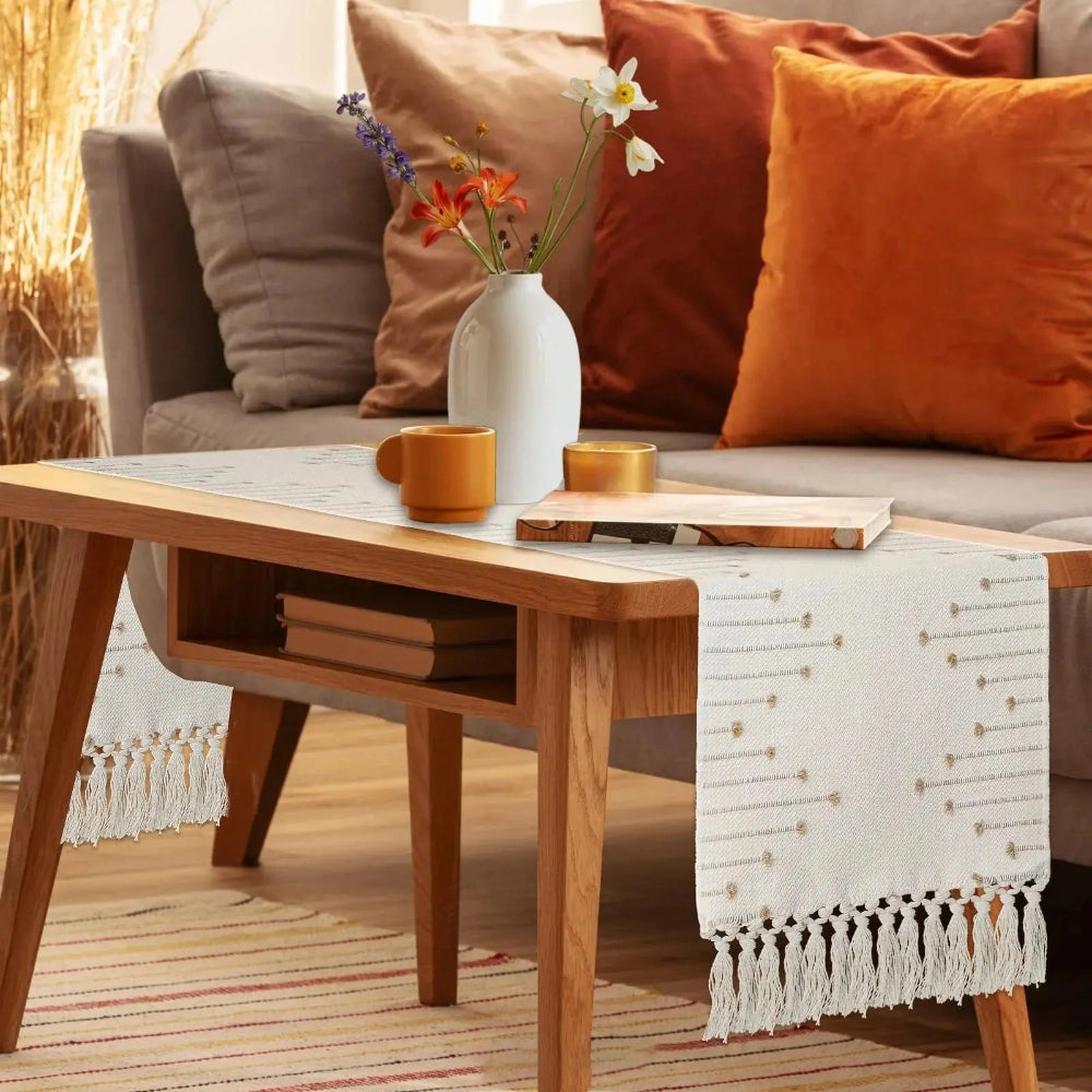 Image d'un chemin de table couleur crème sur une table basse en bois ornée d'un vase de fleurs et d'un livre avec de magnifiques coussins colorés en arrière-plan