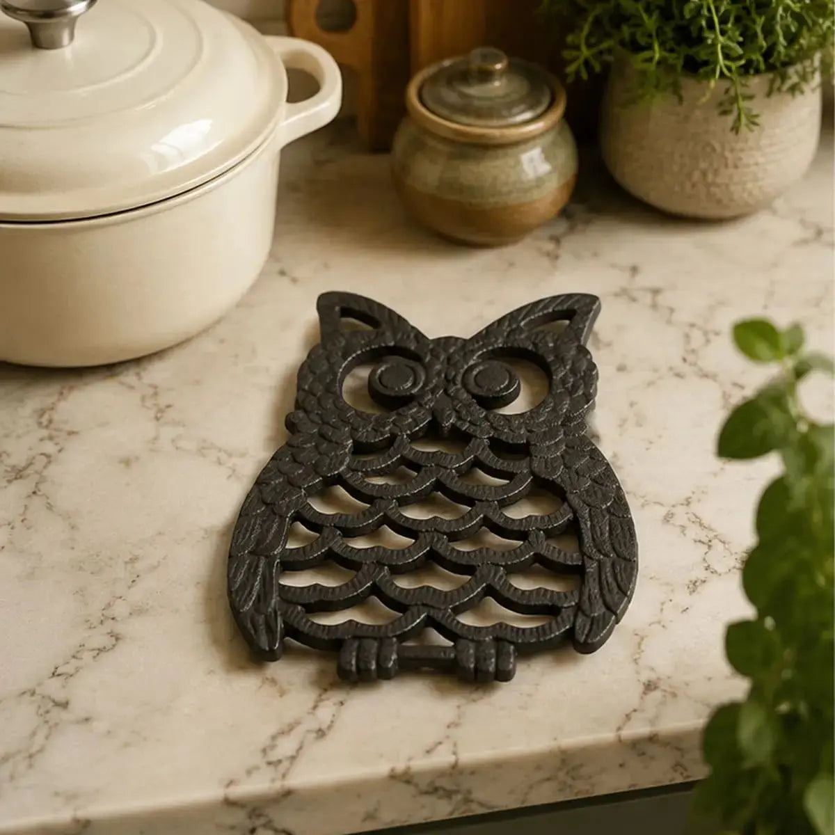 Dessous de plat en fonte en forme de hibou noir posé sur un comptoir en marbre près d'une cocotte en céramique