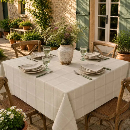 Nappe de table beige à carreaux sur une table carrée en terrasse, entourée de volets verts et de fleurs sauvages