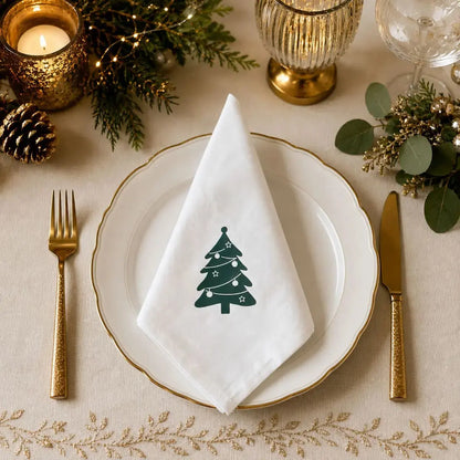 Serviette de table de Noël en coton blanc à motif sapin, pliée sur assiette dorée avec décor de Noël